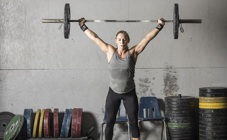 Strong young woman with heavy barbell over her head in grungy gym.