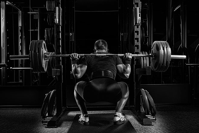 Professional athlete sits with a barbell on his shoulders and prepares to stand with her. View from the back.
